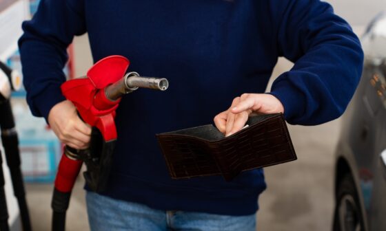 A person holding a fuel nozzle and an empty-looking wallet at a gas station.