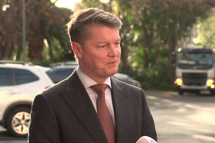 A man with short, fair hair wears a dark suit while standing in a car park.