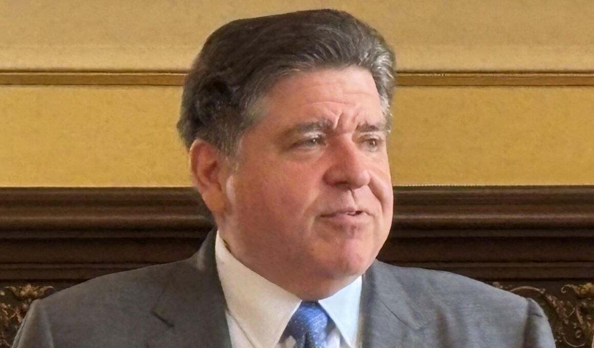 Middle-aged man in a gray suit and blue tie sits in a formal room, looking to the side.